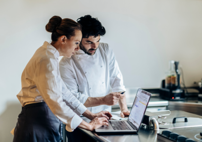 Two restaurant staff members reviewing data on a laptop in a kitchen workspace.